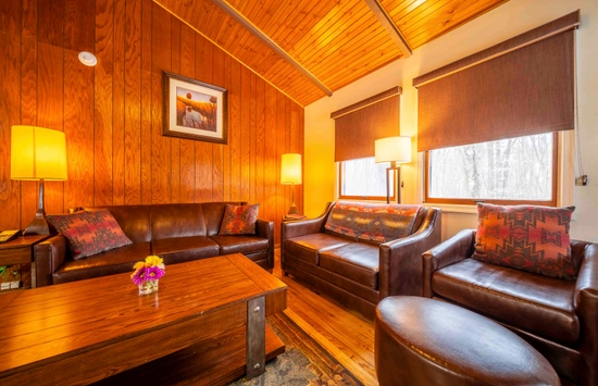 A cozy living room with wood-paneled walls and ceiling, brown leather sofas and chair, two standing lamps, a wooden coffee table with a small flower vase, and two windows with shades letting in soft light.