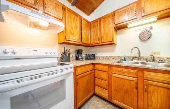 A kitchen with wooden cabinets, a white stove and oven, granite countertops, a double sink, and several appliances including a toaster, coffee maker, and knife block. A light fixture is above the sink.