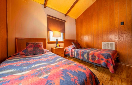 A cozy bedroom with two single beds, each covered in colorful, patterned blankets. A small wooden nightstand with a lamp and flowers sits between the beds. The room features wood-paneled walls and a wood floor.