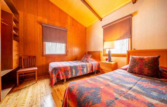 A cozy wooden bedroom with two single beds featuring colorful patterned bedspreads, a nightstand with a lamp and flower vase between them, wooden floors, a chair, and two windows with rolled-down shades.