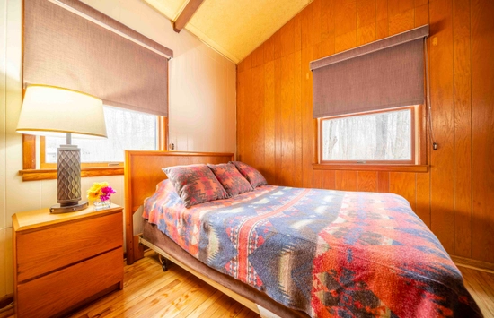 A cozy bedroom with wood-paneled walls, a bed covered in a colorful geometric-patterned blanket, two windows with brown shades, and a wooden nightstand holding a lamp and a small vase of flowers.
