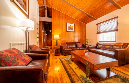 A cozy living room with wood-paneled walls and ceiling, leather sofas, a wooden coffee table, two lamps, framed art, a window with blinds, and a patterned rug on a hardwood floor.