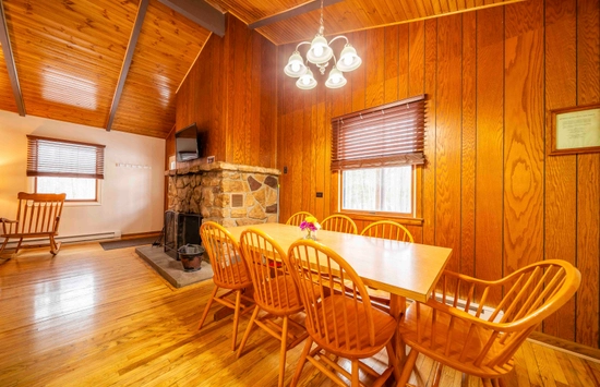 A cozy dining room with wood-paneled walls, a long wooden table surrounded by six matching chairs, a stone fireplace, rocking chair, and a window with blinds letting in natural light.