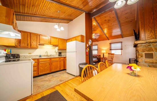 A cozy kitchen and dining area with wooden cabinets, a white refrigerator, a dining table with chairs, and a stone fireplace. The room features wood-paneled ceilings and warm lighting.