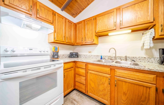 A bright kitchen with wooden cabinets, a white oven and stove, granite countertops, a double sink, and small appliances like a toaster and coffee maker. There are utensils in a holder next to the stove.