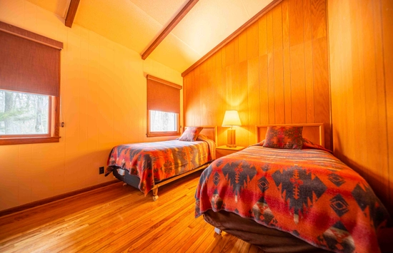 A cozy bedroom with two single beds covered in red and orange patterned blankets, wooden walls, hardwood floor, a lamp on a bedside table, and windows with brown shades letting in natural light.
