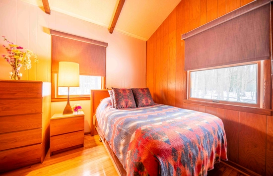 A cozy bedroom with wood-paneled walls, a bed with a colorful patterned blanket, two patterned pillows, a wooden nightstand with a lamp, a dresser with flowers, and large windows with brown shades.