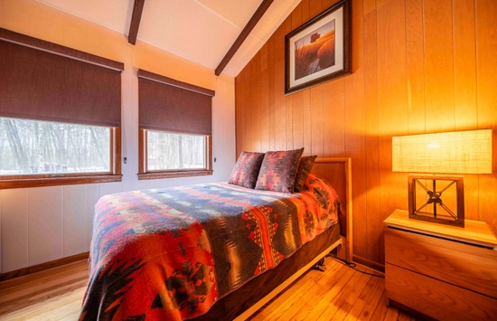 Cozy bedroom with a bed adorned with a colorful Native American-style blanket, two pillows, and a wooden headboard. Two windows offer a view of a snowy forest.