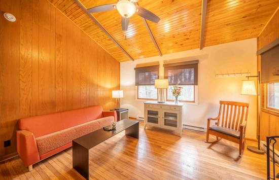Cozy living room with wood-paneled walls and ceiling, a ceiling fan, orange sofa, black coffee table, rocking chair, lamps, and a small cabinet with flowers under two windows. Warm, natural light fills the space.