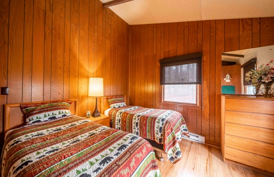 A cozy bedroom with wood-paneled walls, two twin beds with patterned bedding, a lamp between them, a dresser with a mirror, and a window with a dark shade letting in soft light.
