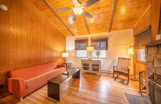 A cozy living room with wood-paneled walls and ceiling, a ceiling fan, a red sofa, a black coffee table, a rocking chair, two lamps, and a stone fireplace. Sunlight streams in through two windows.