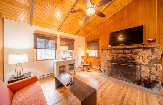 Cozy living room with wood-paneled ceiling, stone fireplace, flat-screen TV, wooden floors, an orange couch, coffee table, rocking chair, and large windows with blinds allowing natural light inside.