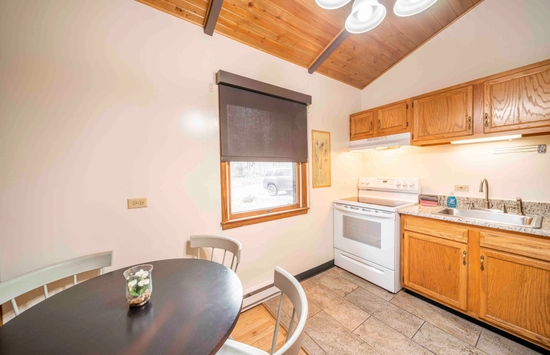 Cozy kitchen with a round dining table and chairs, a window with a black shade, white stove, wooden cabinets, sink, and overhead lighting. The ceiling is wood-paneled, and natural light enters through the window.
