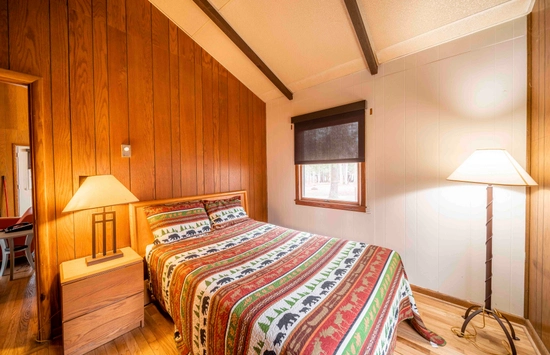 A cozy bedroom with wood-paneled walls, a bed with a colorful patterned quilt, two lamps, a window with a black shade, and hardwood floors. Warm lighting creates a rustic, inviting atmosphere.