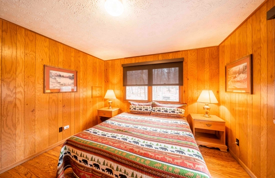 A cozy bedroom with wood-paneled walls, a bed covered in a colorful wildlife-themed quilt, two bedside tables with lamps, and paintings on the walls. Large windows let in natural light and show a snowy scene outside.