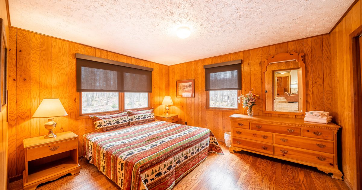 A cozy bedroom with wood-paneled walls and floor, a large bed with a colorful patterned quilt, two nightstands with lamps, a dresser with a mirror, and windows with dark shades letting in natural light.