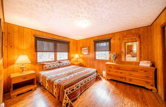 A cozy bedroom with wood-paneled walls, a large bed with patterned bedding, two bedside tables with lamps, a dresser with a mirror, and windows letting in natural light.