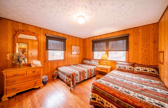 A cozy bedroom with two beds covered in colorful, patterned quilts, wood-paneled walls, a wooden dresser with a mirror and flowers, and large windows with black blinds letting in natural light.