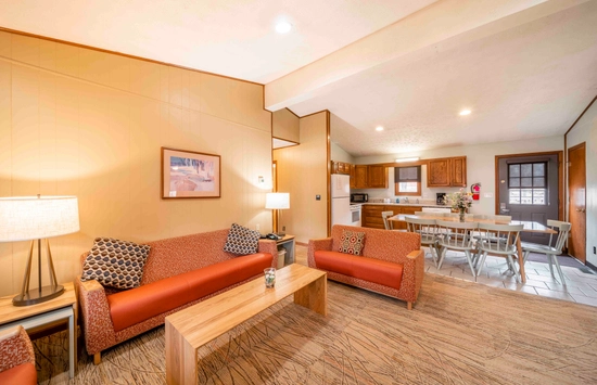 A cozy living room with two orange sofas, a wooden coffee table, lamps, and wall art. The space extends to an open kitchen with wooden cabinets, a dining table, and tile flooring. Warm lighting creates a welcoming atmosphere.