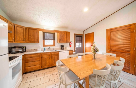Bright kitchen with wooden cabinets, white appliances, and a large wooden table surrounded by six white chairs. A vase with flowers sits on the table. The room has tiled floors and wood-paneled doors.