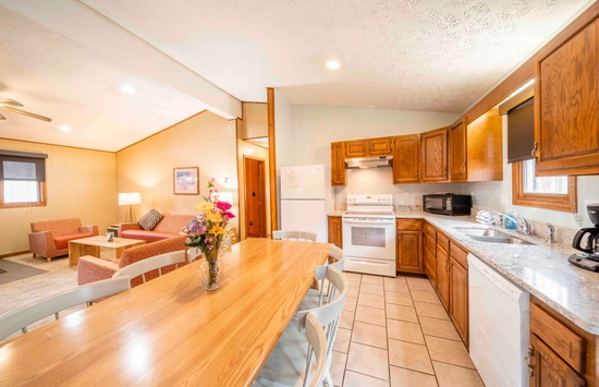 A bright, cozy kitchen with wooden cabinets, white appliances, and a long dining table with a flower vase. In the background, there’s a living area with sofas, a rug, and a lit lamp.