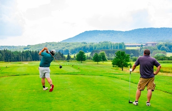 Two men are on a lush green golf course. One is swinging a golf club, while the other stands watching with a club in hand. Trees and hills are visible in the scenic background under a partly cloudy sky.