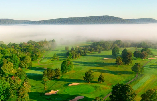 Aerial view of a green golf course with sand bunkers and trees, surrounded by rolling hills. Morning fog blankets the background, partially covering distant wooded hills under a clear sky.