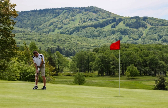 A man is putting on a golf green near a red flag, with lush green trees and a large forested mountain in the background under a partly cloudy sky.