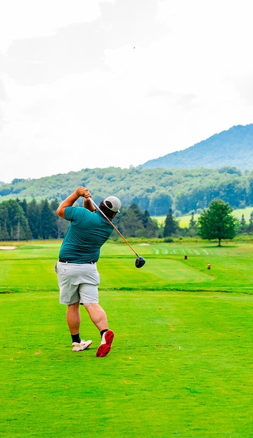 A golfer wearing a cap, teal shirt, and gray shorts swings a golf club on a green course, with trees and a mountain visible in the background.