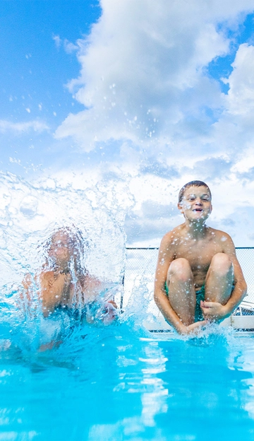 Two children are jumping into a bright blue swimming pool, creating a large splash, with blue sky and clouds visible in the background. One child is clearly seen mid-air while the other is mostly obscured by water.