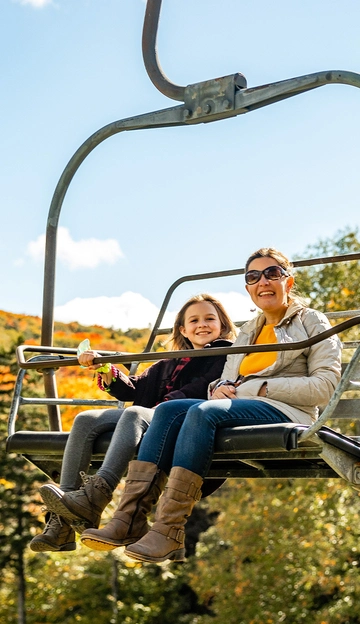 A woman and a young girl, both smiling, sit together on a chairlift outdoors. They're dressed in jackets and boots, surrounded by trees with autumn foliage under a bright blue sky.