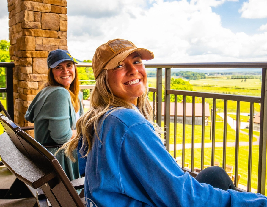 Two women in casual clothing and hats sit smiling on chairs on a balcony, with green fields and a cloudy sky visible in the background. One woman looks at the camera, while the other looks to the side.