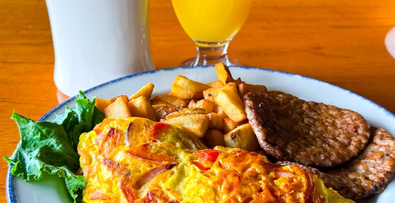 A plate with a vegetable omelet, breakfast potatoes, sausage patties, and lettuce, with a cup of coffee and a glass of orange juice on a wooden table.
