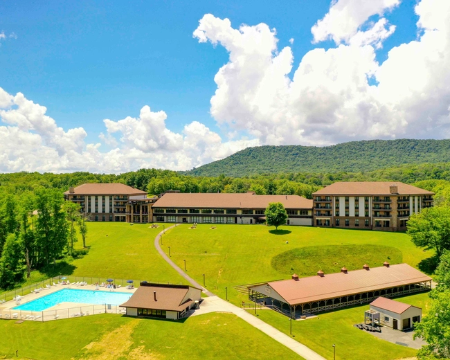 A large resort complex with three connected buildings sits on a green lawn, surrounded by trees and mountains. A swimming pool and pavilion are visible in the foreground under a blue sky with fluffy clouds.