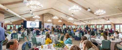 A large group of people attend a banquet or conference in a spacious, well-lit hall with round tables, decorated centerpieces, and a presentation screen at the front. Some attendees are listening while others talk among themselves.