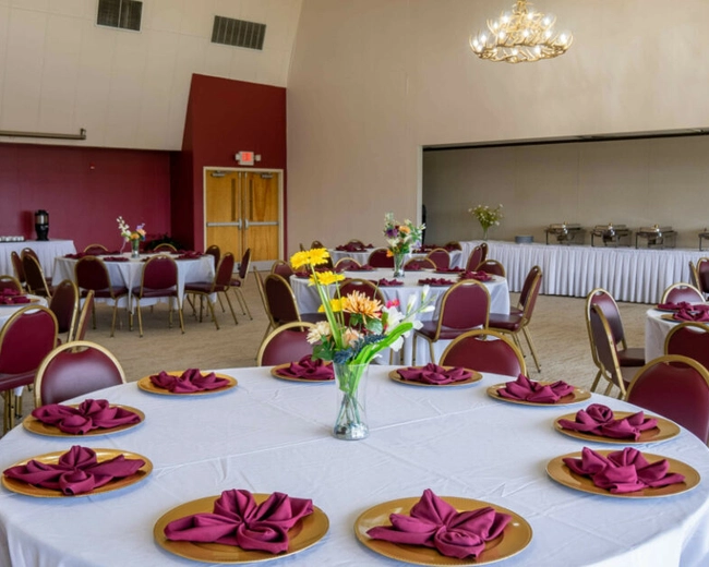 A banquet hall with round tables covered in white tablecloths, set with burgundy napkins and gold plates. Each table has a vase of flowers, and buffet tables line the back wall. Burgundy chairs surround the tables.