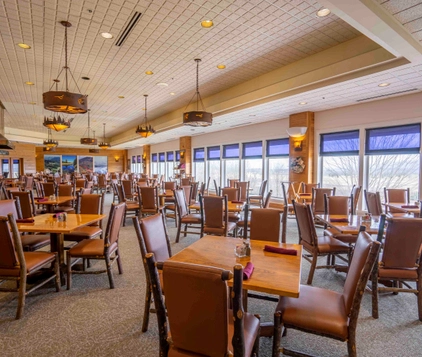 Spacious restaurant dining area with many wooden tables and chairs, large windows letting in natural light, and scenic views outside. The room is empty and warmly lit with hanging lamps.