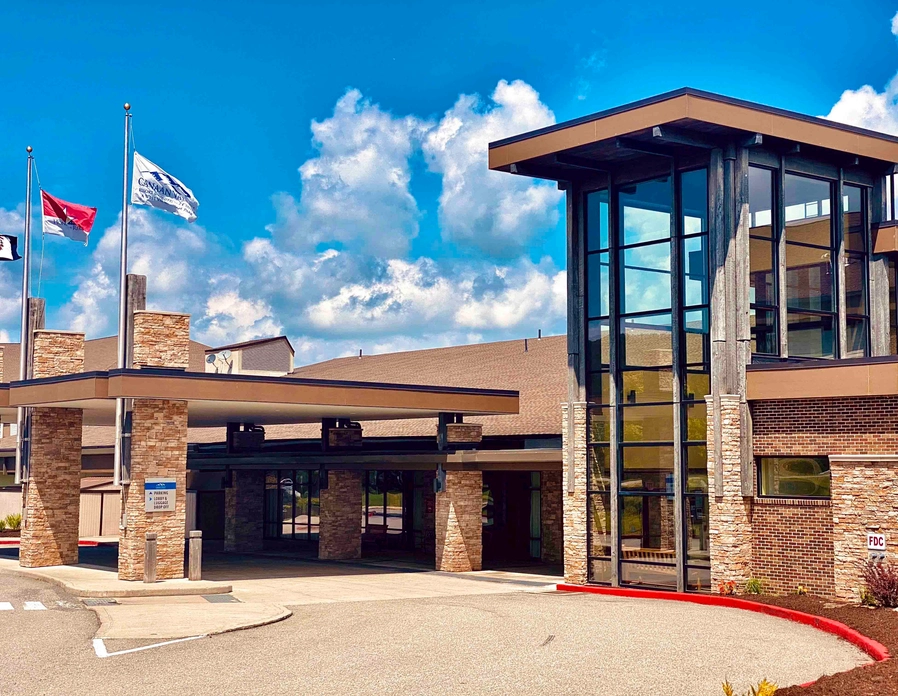Modern building entrance with large glass windows, stone pillars, and a covered drop-off area. Three flags are flying on poles outside, and the sky is bright with scattered clouds.