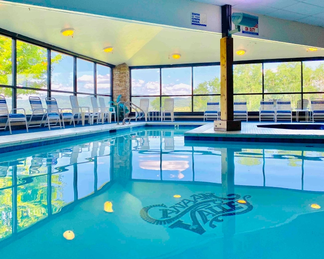 Indoor swimming pool with clear blue water, surrounded by white chairs and large floor-to-ceiling windows letting in bright natural light and offering views of green trees outside. The pool has a logo on its floor.