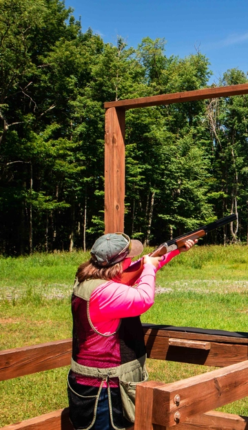 A person wearing a cap and pink shirt aims a shotgun upward at an outdoor shooting range, surrounded by green grass and trees under a blue sky.