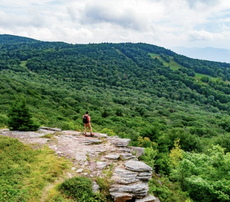 A person with a backpack stands on a rocky outcrop overlooking a lush, green forested valley and distant rolling hills under a partly cloudy sky.