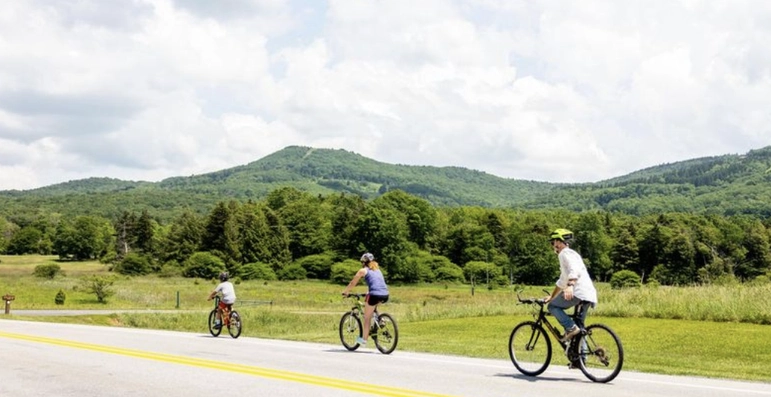 Three people ride bicycles along a rural road with green grass and trees on both sides and forested hills in the background under a partly cloudy sky.