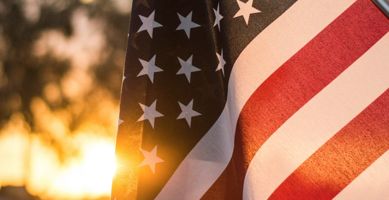 Close-up of the United States flag with stars and stripes, illuminated by warm sunlight during sunset, with a blurred outdoor background.