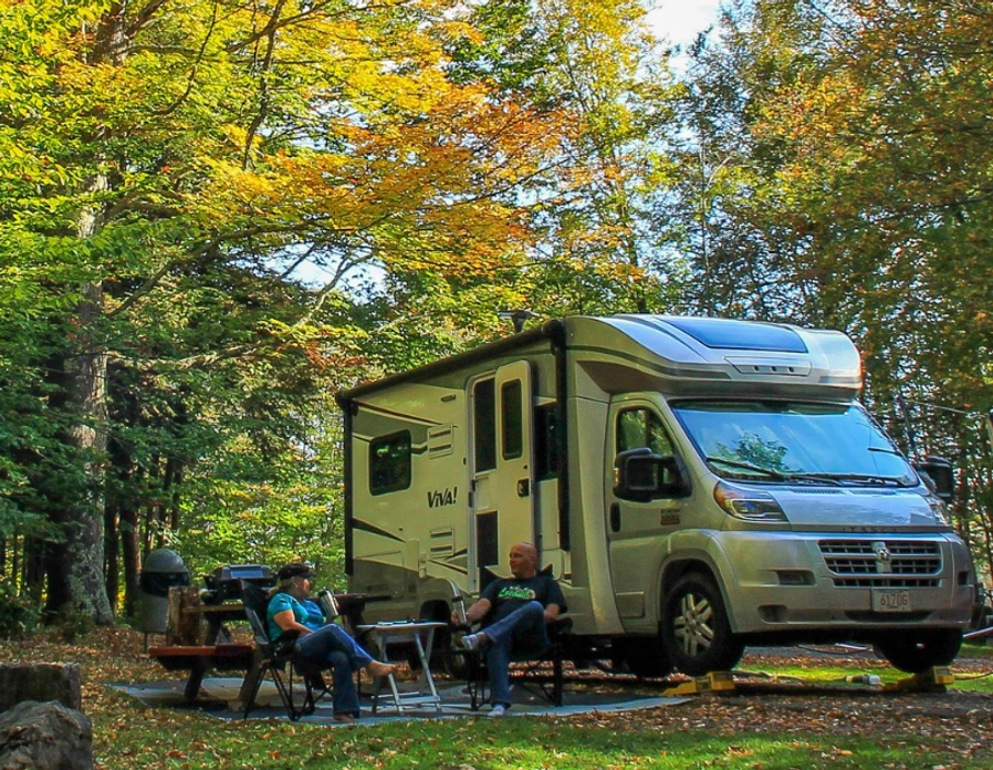 A silver RV is parked in a wooded campsite. An American flag hangs from a clothesline. Two people sit in chairs, enjoying the autumn scenery.