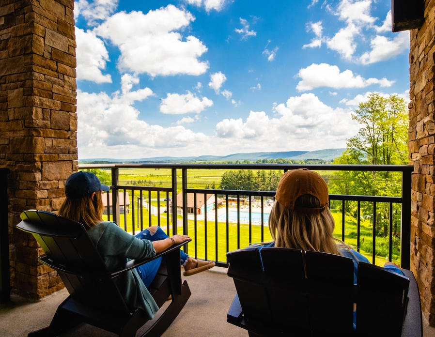 Two people in hats sit in wooden chairs on a balcony, looking out over a scenic view of green fields, trees, mountains, and a pool under a bright blue sky with fluffy clouds.