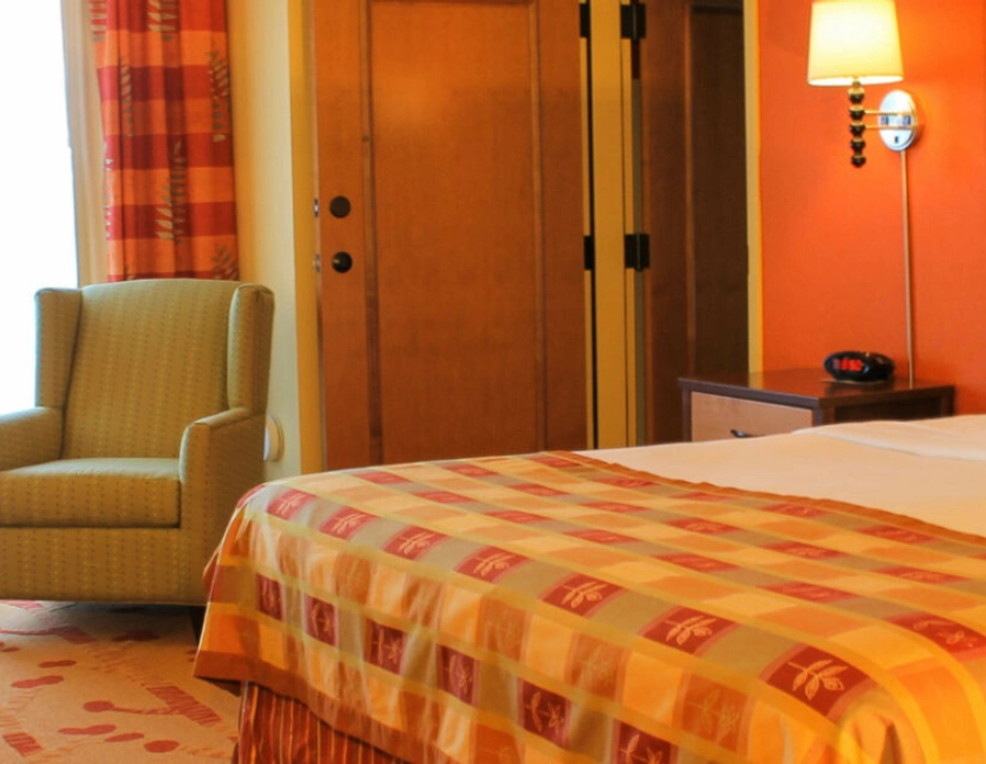 A hotel room with a large bed, four white pillows, a patterned bedspread, an armchair, a microwave, a mini-fridge, a desk, and bright natural light coming through a window with red curtains.