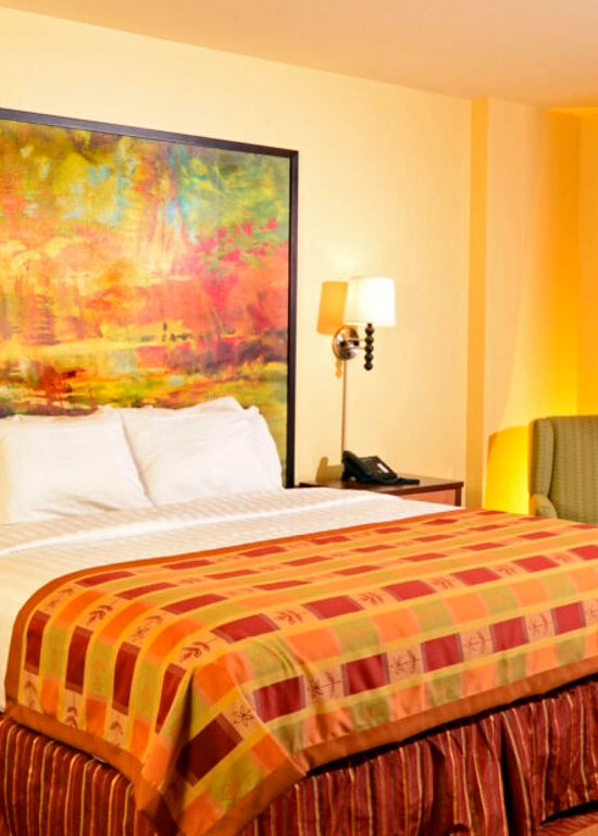 A bright hotel room with a king bed, colorful bedspread, vivid abstract painting above the headboard, nightstands with lamps, a green armchair by the window, and glass doors leading to a balcony with a view.