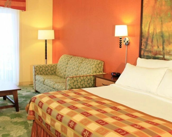 A bright hotel room with a large bed, white pillows, patterned bedspread, a green sofa, wooden coffee table, two lamps, floral carpet, and a window with sheer curtains and red drapes.