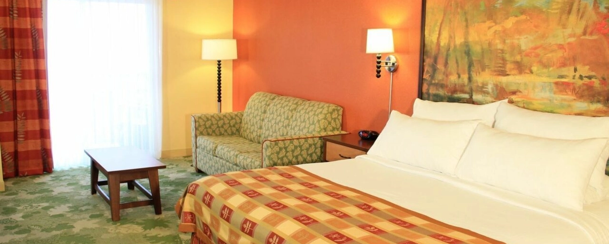 A hotel room with a king bed, patterned bedspread, green sofa, wooden coffee table, two lamps, large window with sheer curtains, and a colorful abstract painting above the bed.