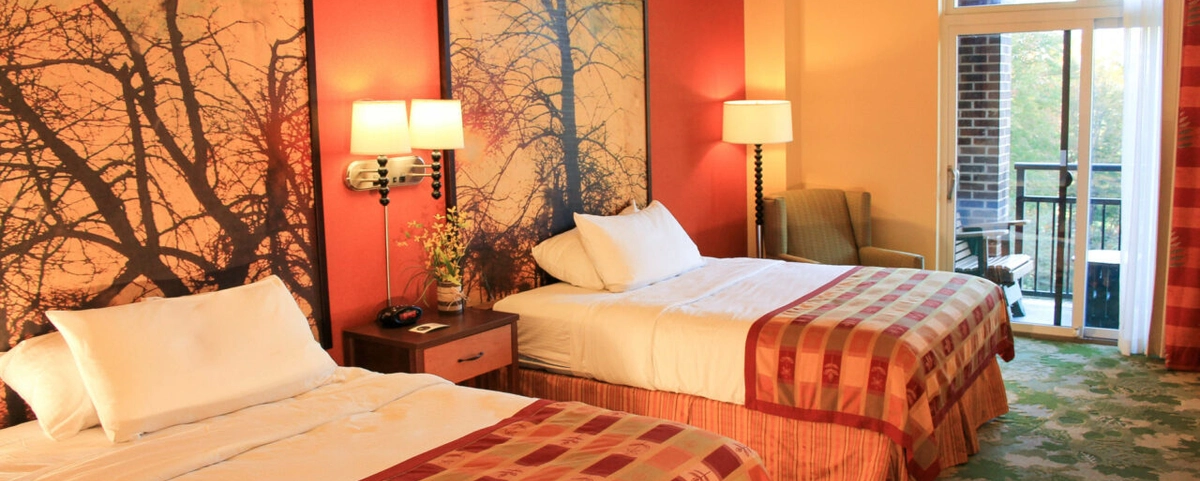 A hotel room with two double beds, each with red patterned bedding, a nightstand with a phone and lamp between them, wall art of tree silhouettes, an armchair, and a glass door leading to a balcony.
